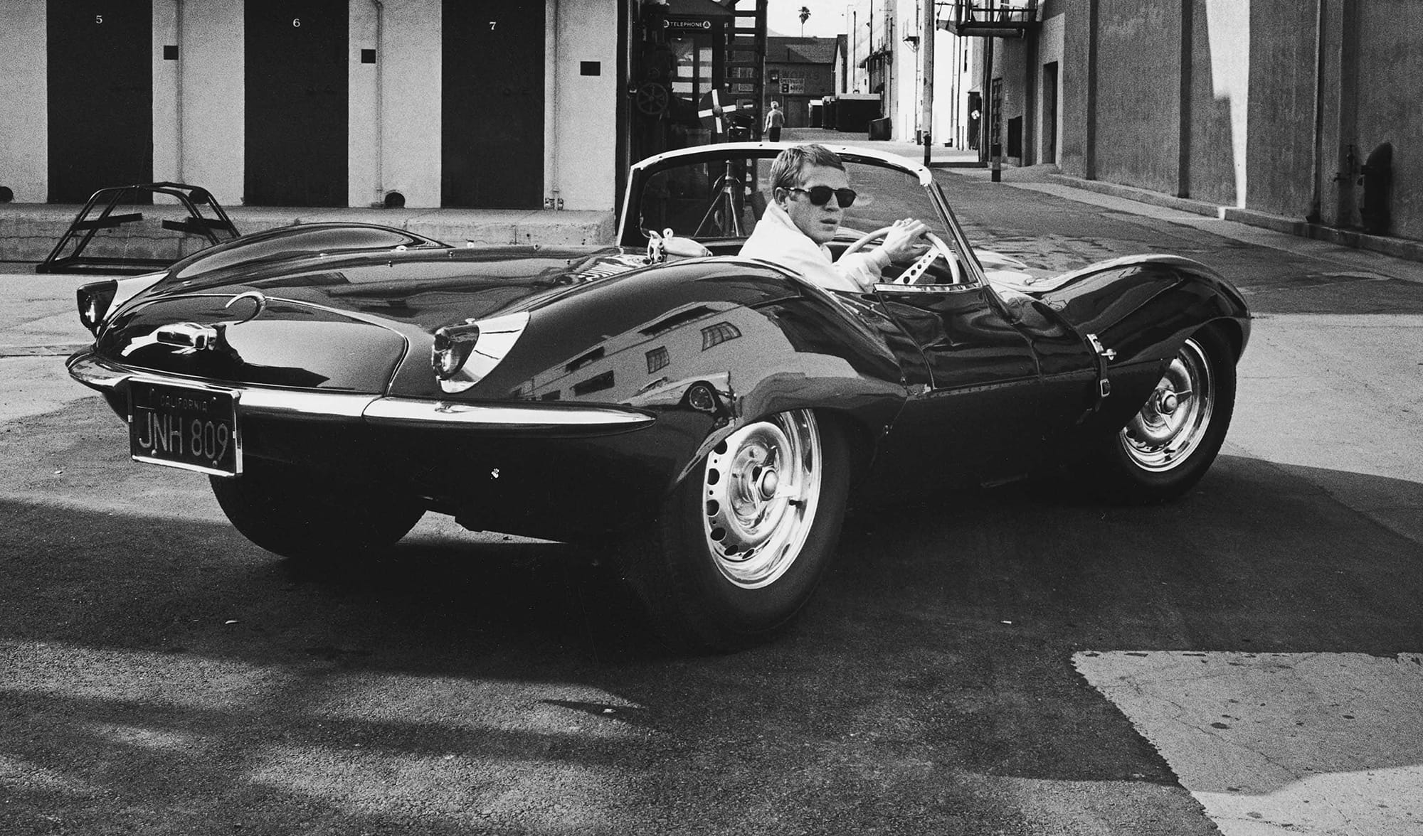 Man driving a vintage sports car on a city street, wearing sunglasses and a white shirt, with buildings in the background.