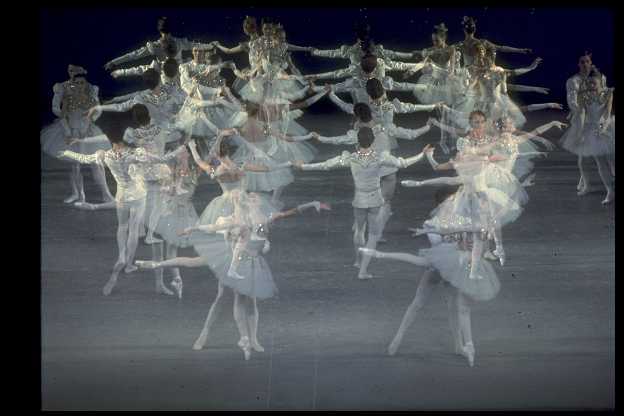 Ballet dancers in synchronized formation, wearing white costumes, performing on stage with a dark background.