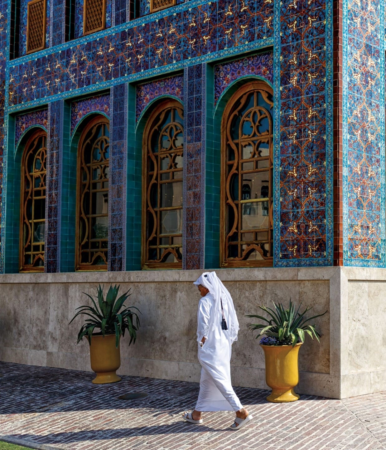 Person walking by colorful, ornate building with arched windows and potted plants on a sunny day.