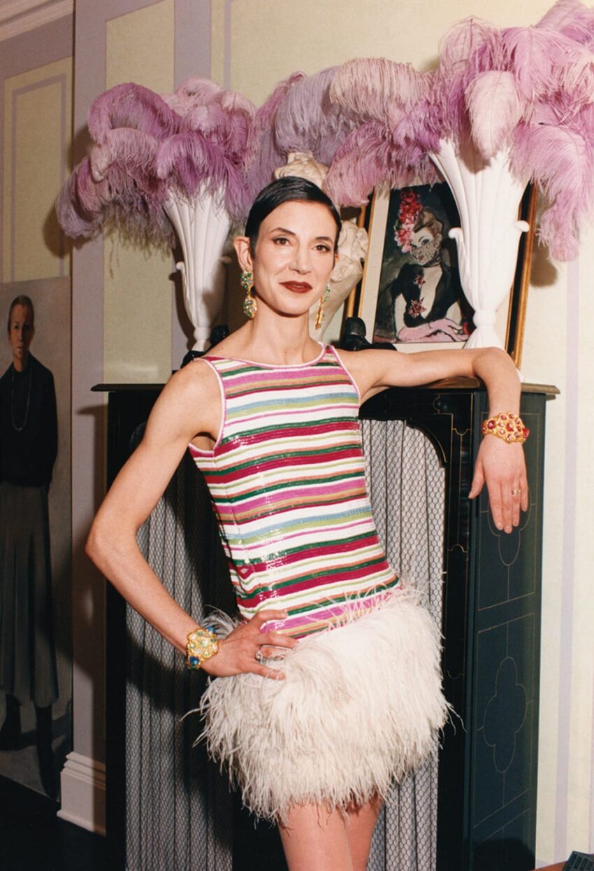 Person in a striped top with a feathered skirt, standing beside a mantel with decorative feathers and a portrait.