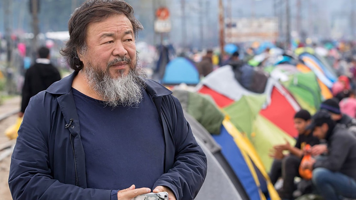 Man with a beard stands near a group of colorful tents on a cloudy day.