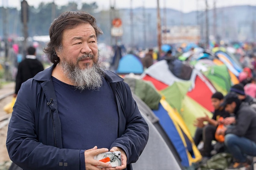 Man with a beard stands near a group of colorful tents on a cloudy day.
