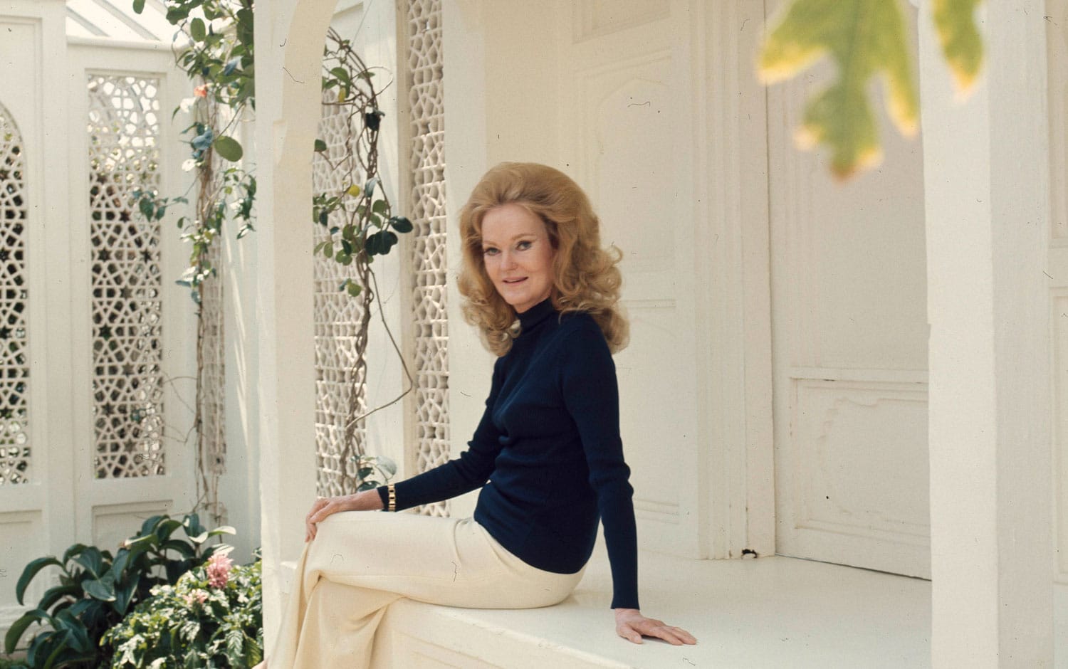 A woman with long hair in a blue sweater and white pants sits on a white porch surrounded by plants.
