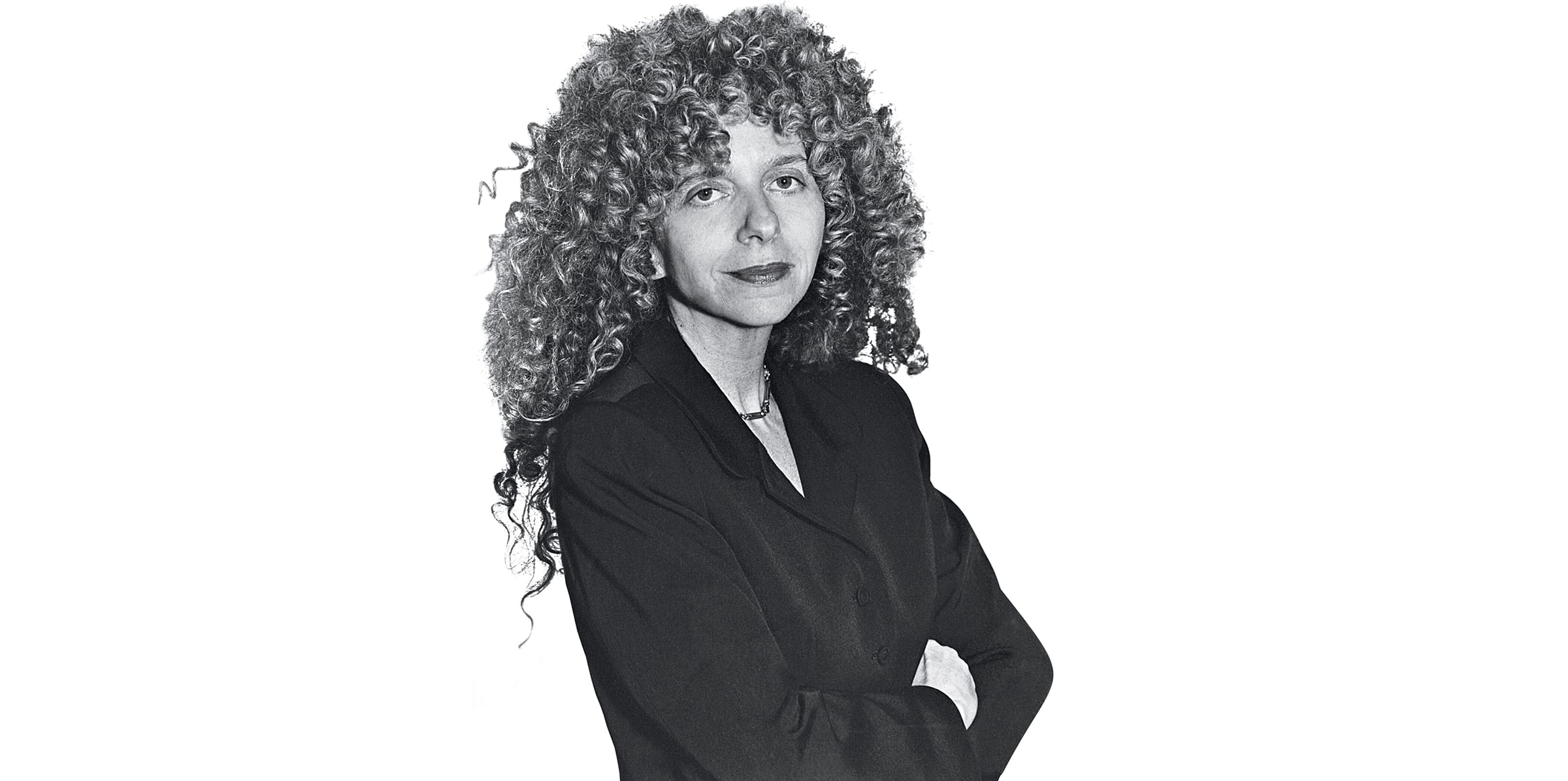 Person with long curly hair wearing a dark blazer, arms crossed, looking at the camera in a black and white photo.
