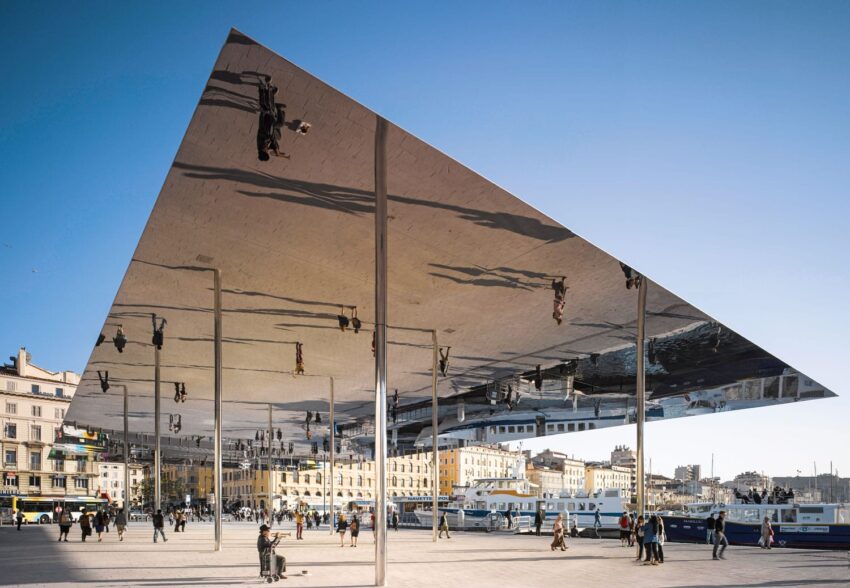 Modern mirrored pavilion with reflections above a sunny public square, with people walking and historic buildings in the background.