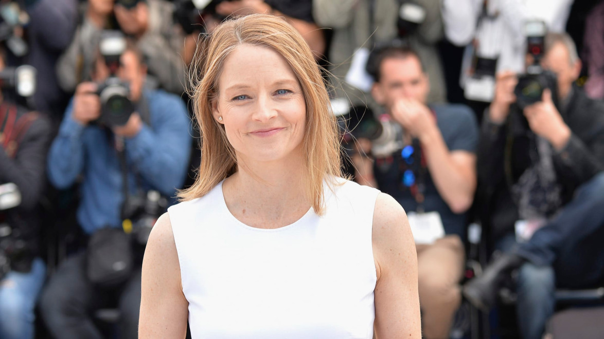 Person smiling at a public event with photographers in the background, wearing a white top and standing outdoors.