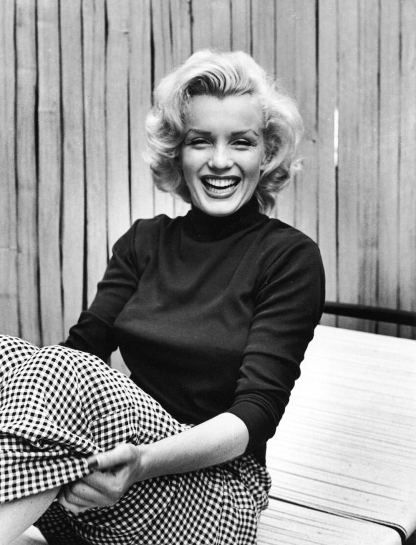 Person smiling, sitting outdoors in a checkered skirt and dark top with short, wavy hair against a wooden backdrop.