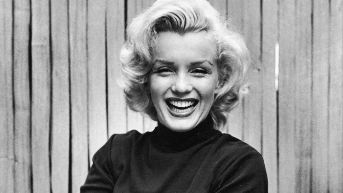 Smiling woman with blonde hair wearing a dark top against a wooden backdrop
