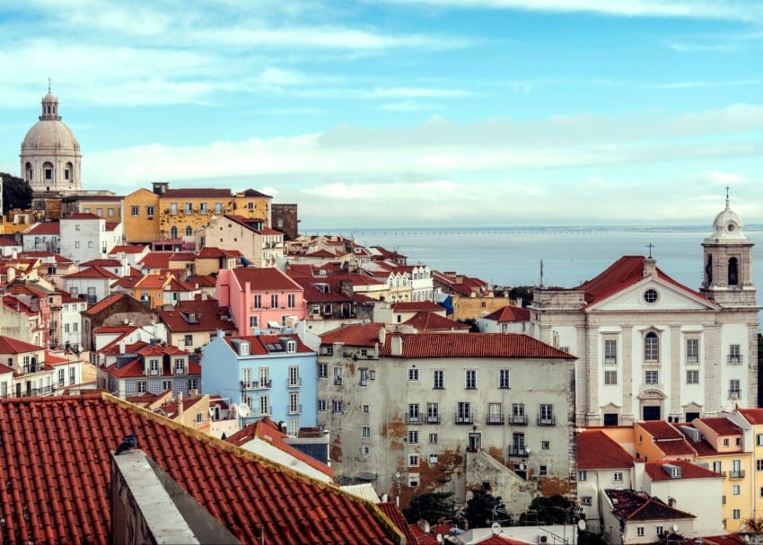 Colorful rooftops and historic buildings under a blue sky in Lisbon, Portugal, with the Tagus River in the background.