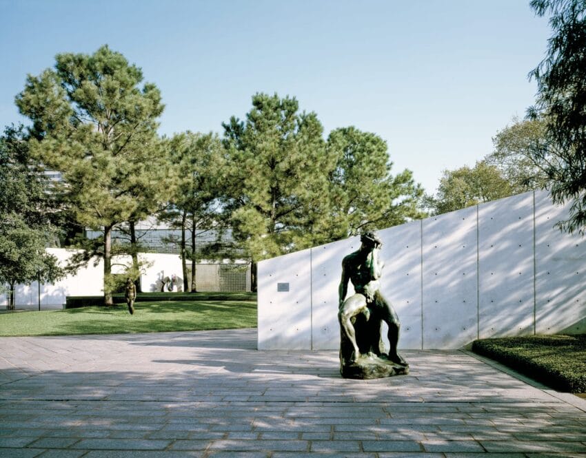 Sculpture of a seated figure in a sunlit outdoor courtyard surrounded by trees and modern architecture.