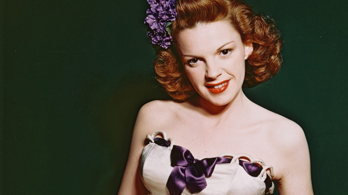 Person with vintage hairstyle and floral hair accessory, wearing a strapless dress with purple ribbon, smiling against dark background.