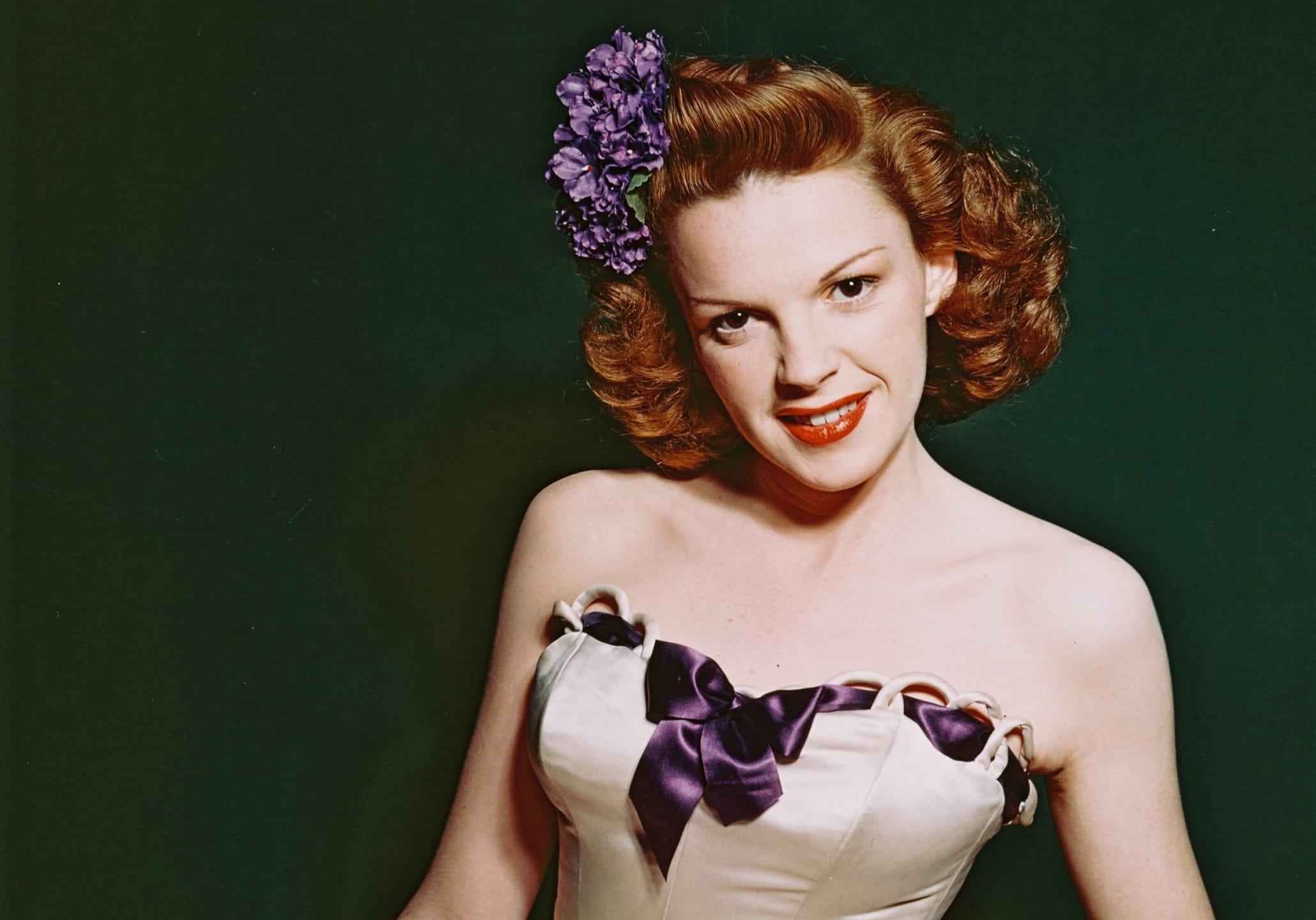 Person with vintage hairstyle and floral hair accessory, wearing a strapless dress with purple ribbon, smiling against dark background.