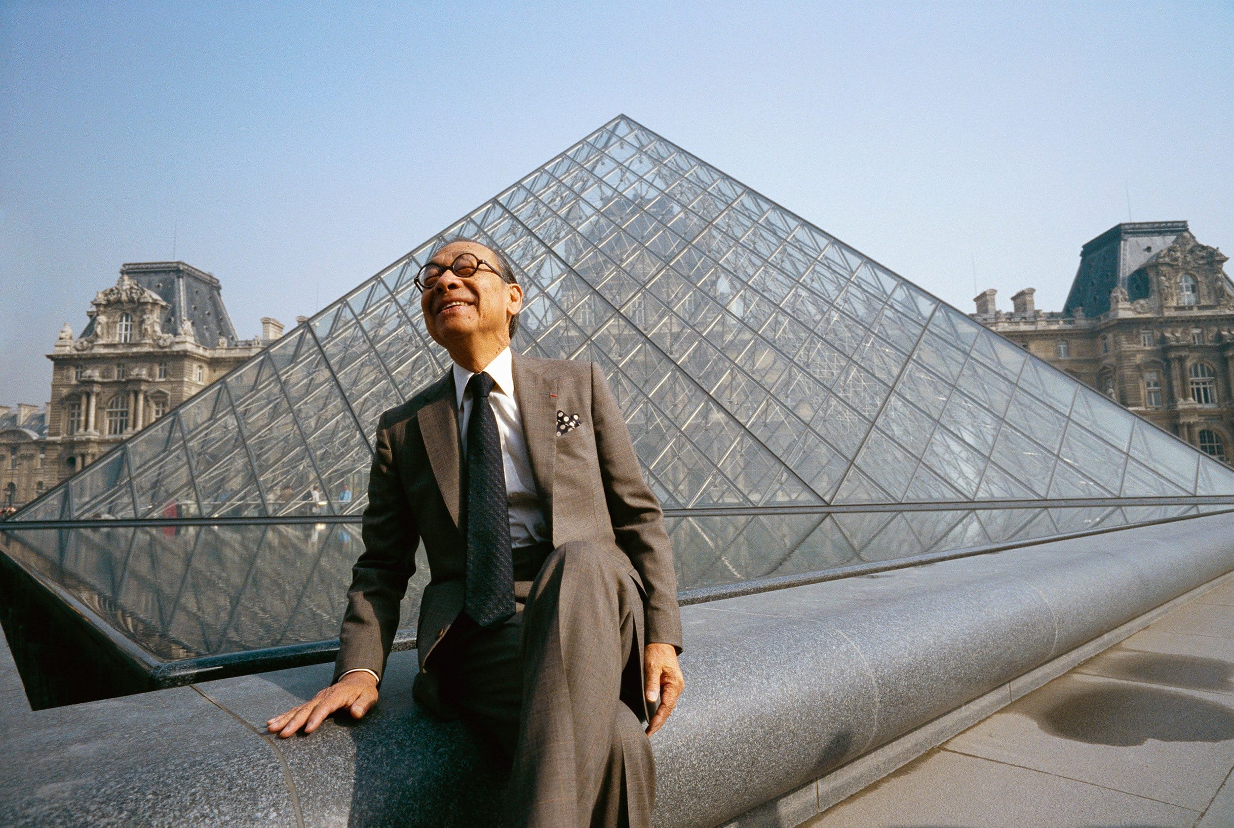Person in a suit sitting in front of the Louvre Pyramid on a sunny day.
