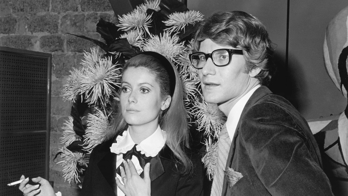 A black and white photo of a woman with a cigarette and a man with glasses, both dressed in formal attire.