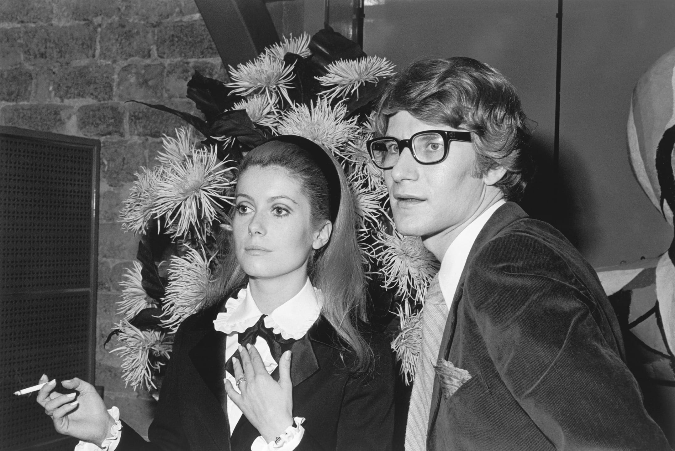 A black and white photo of a woman with a cigarette and a man with glasses, both dressed in formal attire.