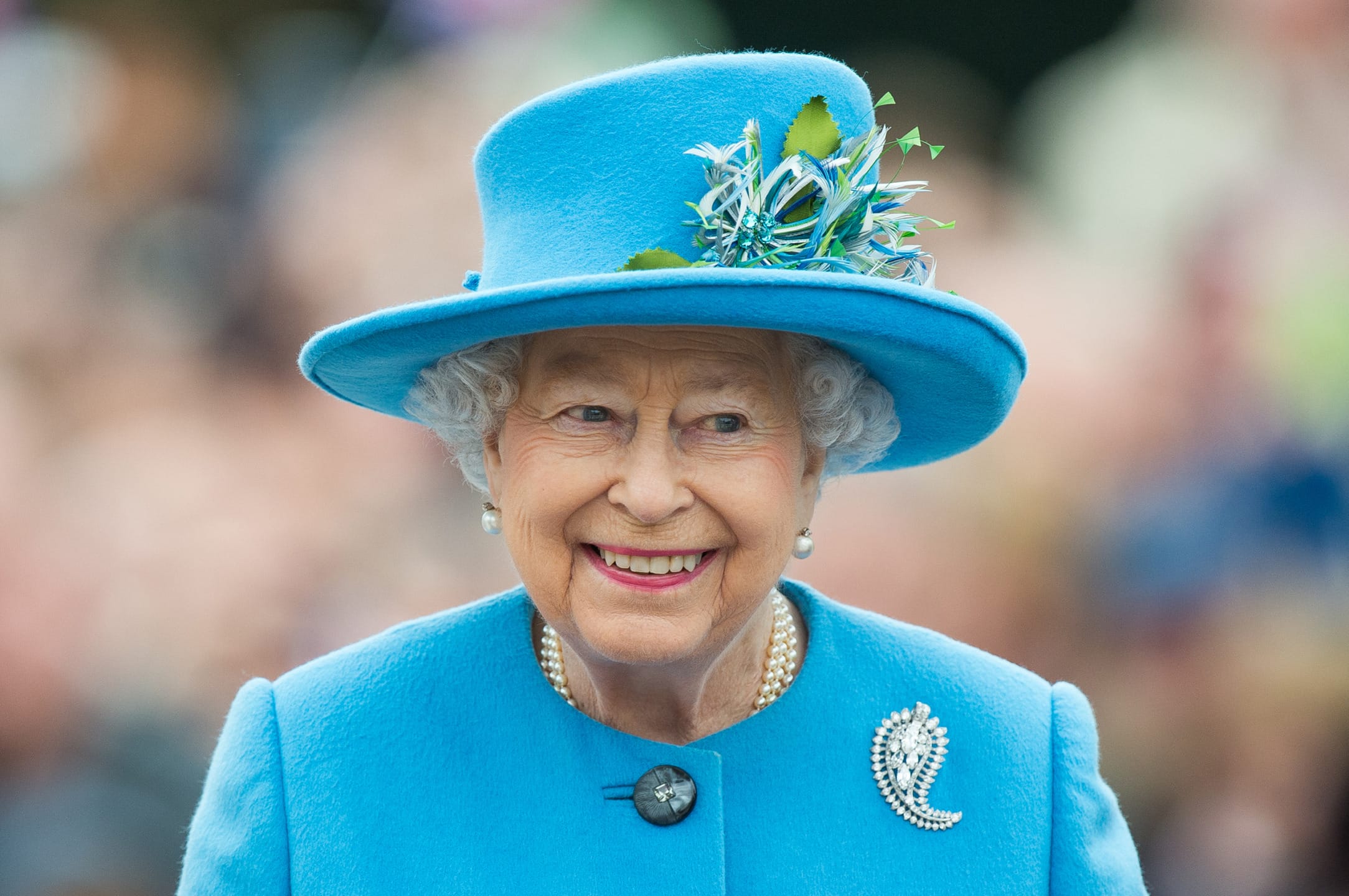Elderly woman in bright blue outfit and hat with pearls and a flower accessory, smiling outdoors.