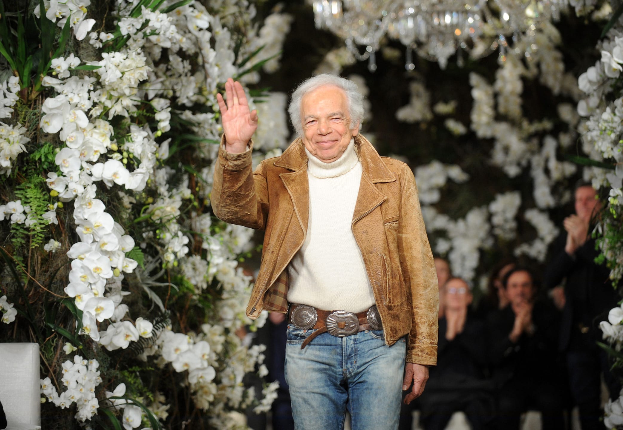 Person in a brown suede jacket and jeans waves while surrounded by white flowers and greenery.