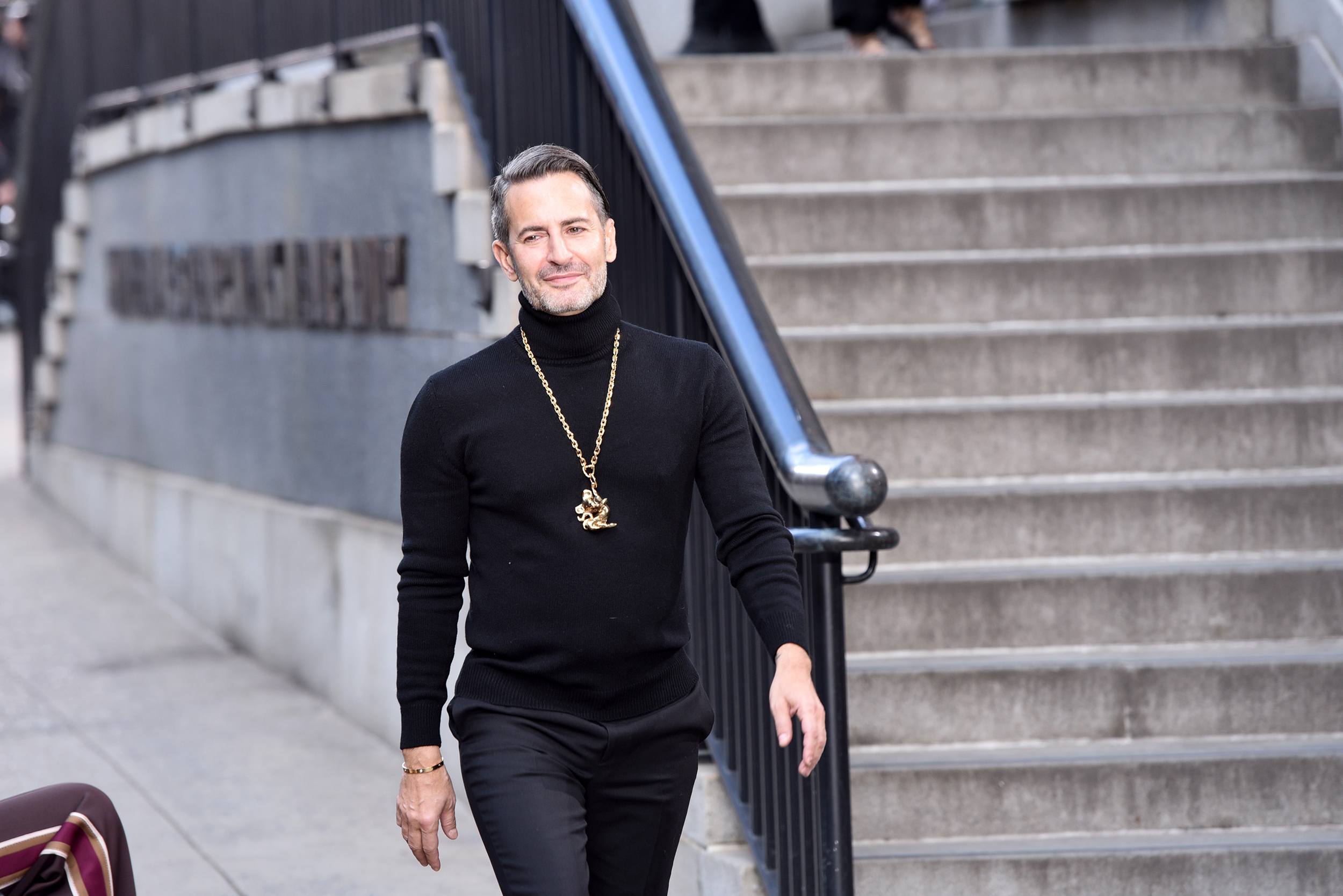 Person in black turtleneck and gold necklace walking down stairs beside a railing.