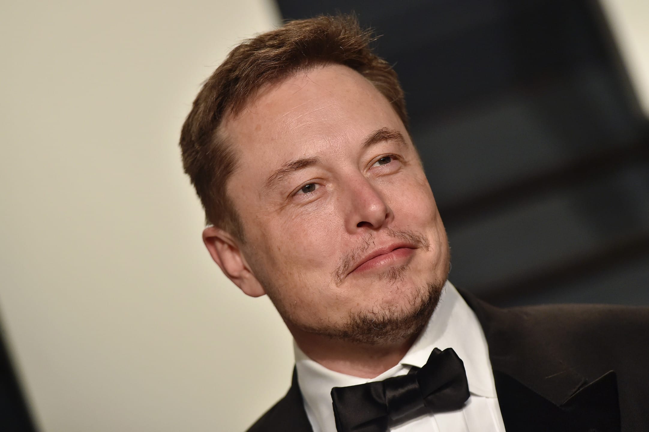 Man in formal attire with a bow tie, smiling slightly, standing against a blurred background.