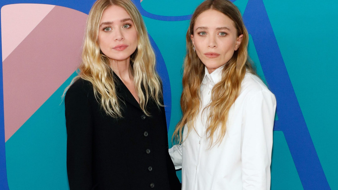 Two individuals posing together in stylish outfits against a colorful abstract background.