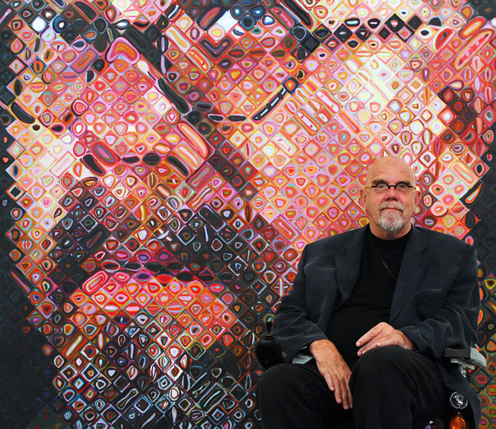 Man seated in front of a large, colorful abstract portrait with geometric patterns.