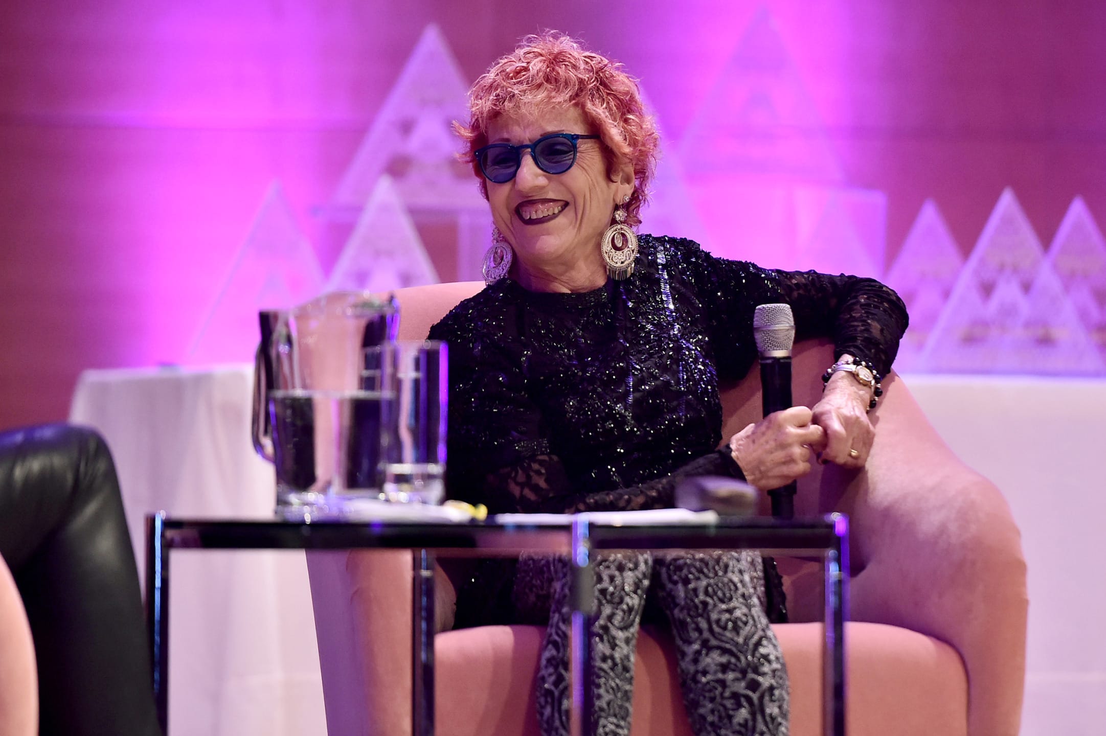 Person sitting in a pink chair on stage, holding a microphone and smiling, against a pink and purple backdrop.