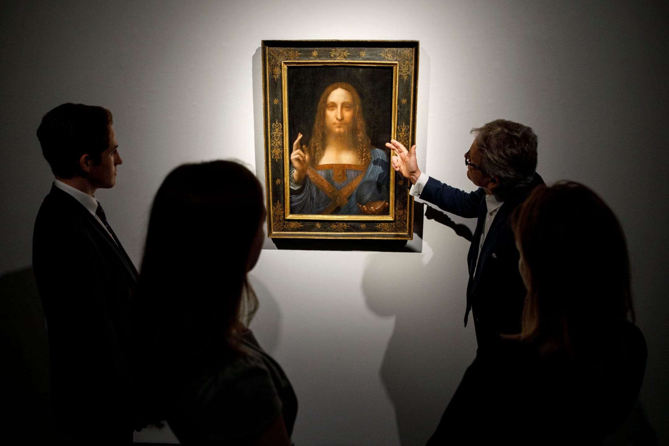 People observing a framed painting of Jesus, with one person pointing at the artwork, in a dimly lit gallery setting.