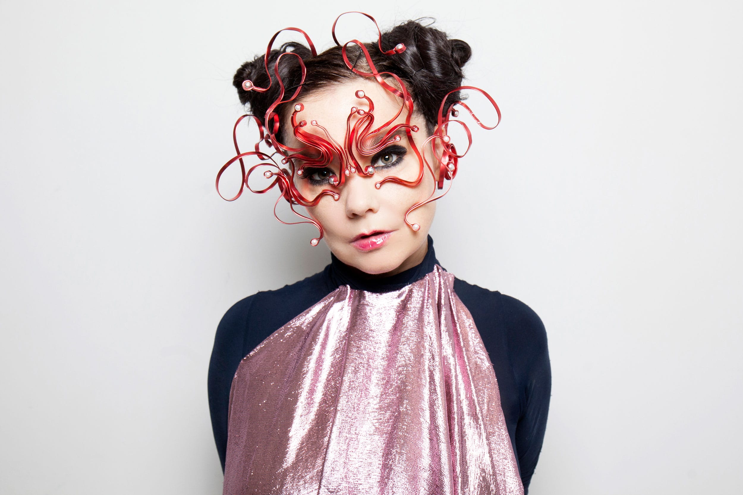 Person wearing a unique red artistic mask with curled designs and a shiny pink outfit against a plain background.
