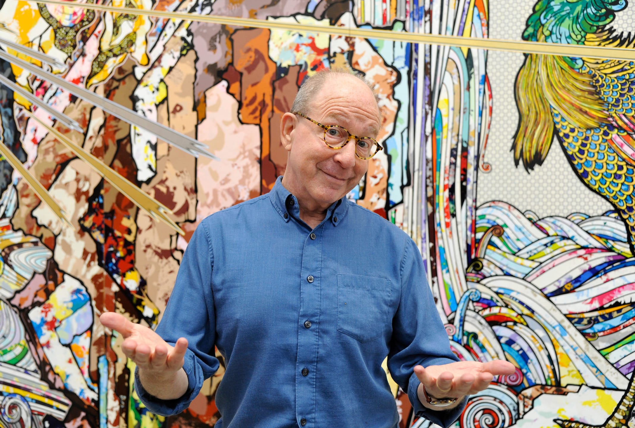 Man in blue shirt with glasses stands smiling in front of colorful, abstract mural with diverse patterns and textures.
