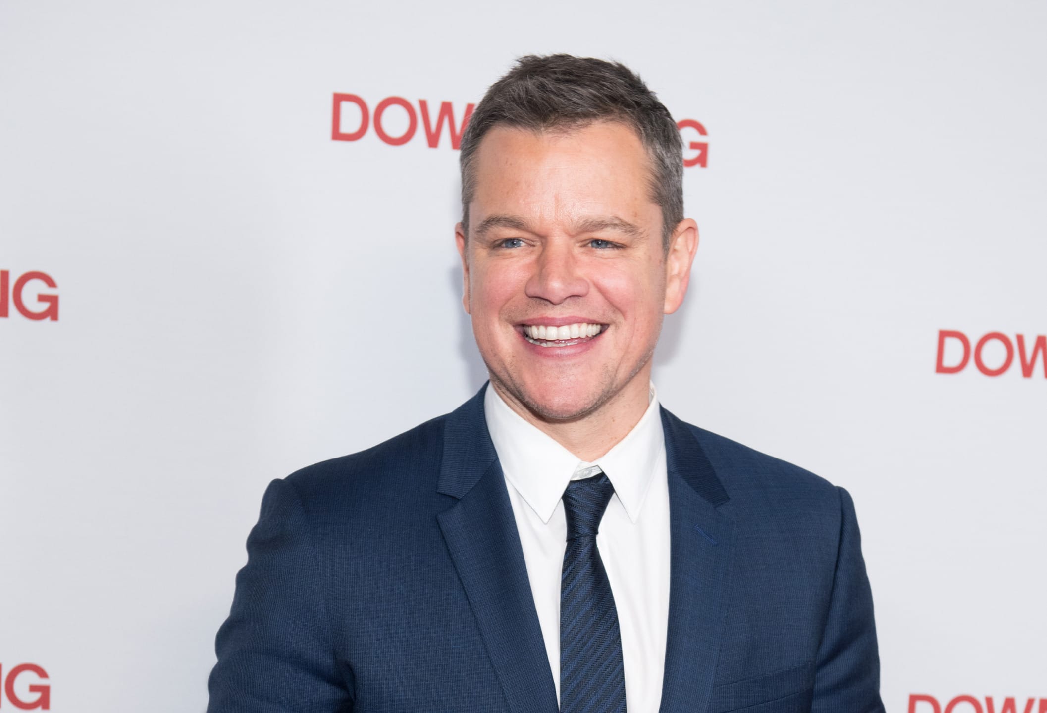 Man in a blue suit smiling in front of a wall with red letters.