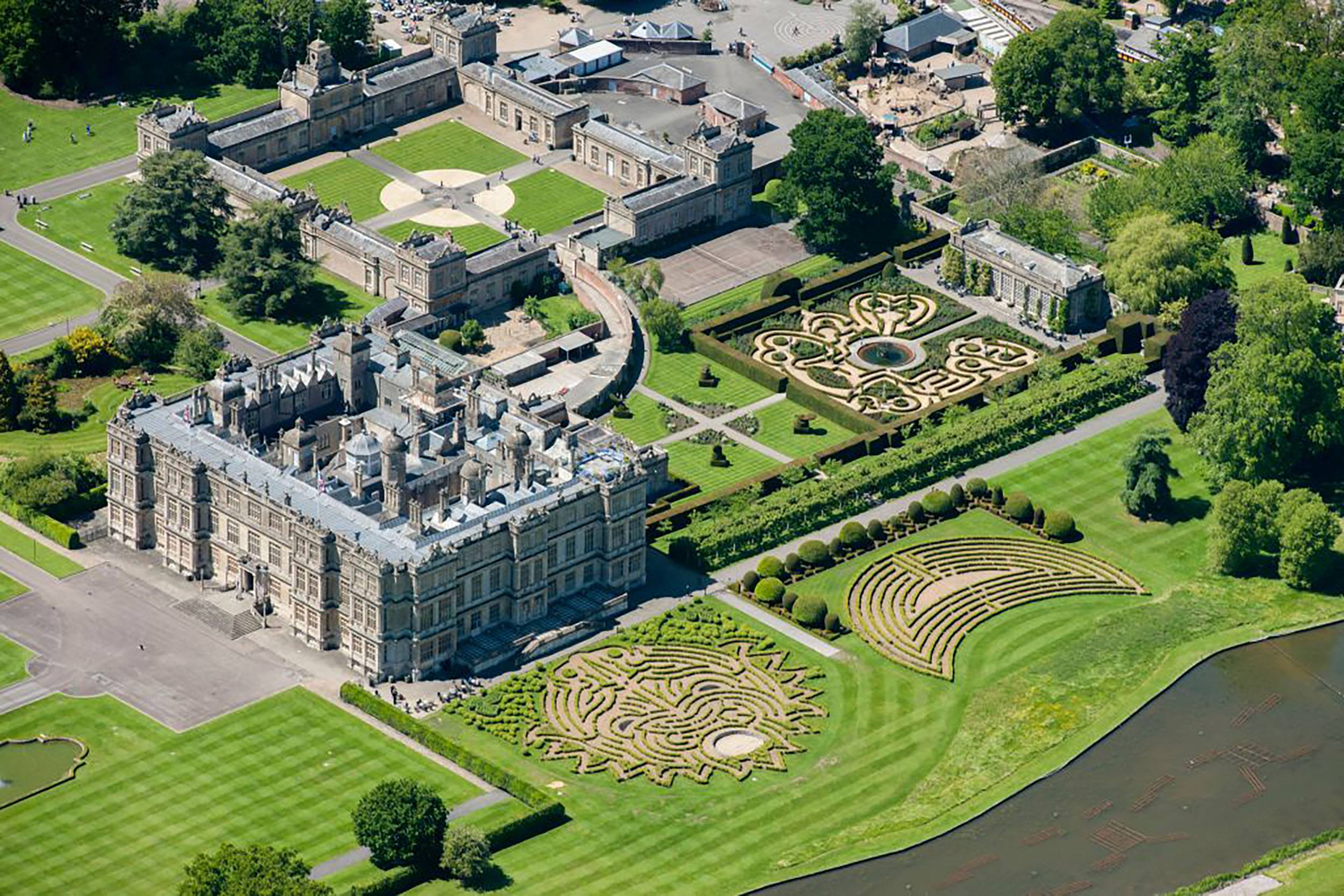 Aerial view of a large historic mansion with intricate gardens and symmetrical pathways surrounded by lush greenery.