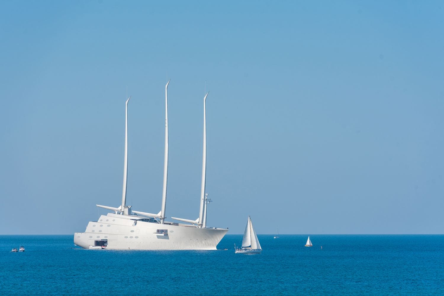 A large, modern sail yacht with three masts on a calm blue sea, surrounded by smaller sailboats on a clear day.