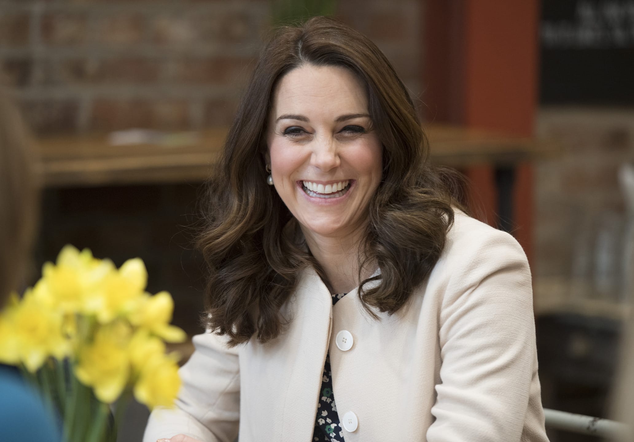Smiling woman with long brown hair sitting at a table with a bouquet of yellow flowers in the foreground.