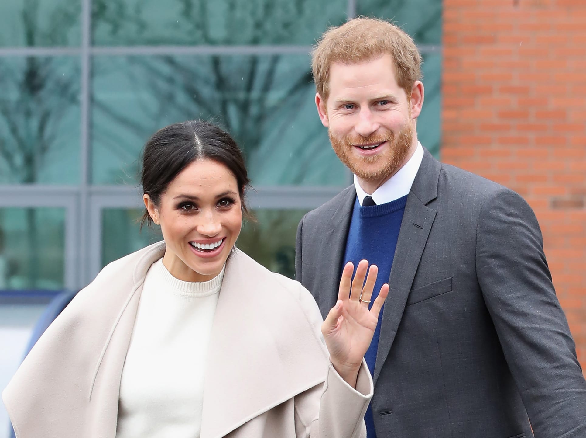 A smiling woman and man in formal attire waving outdoors in front of a building.