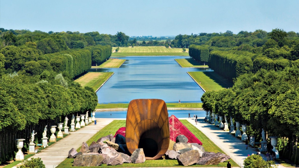 Sculpture in a garden with a long canal, surrounded by trees and statues, under a clear blue sky.