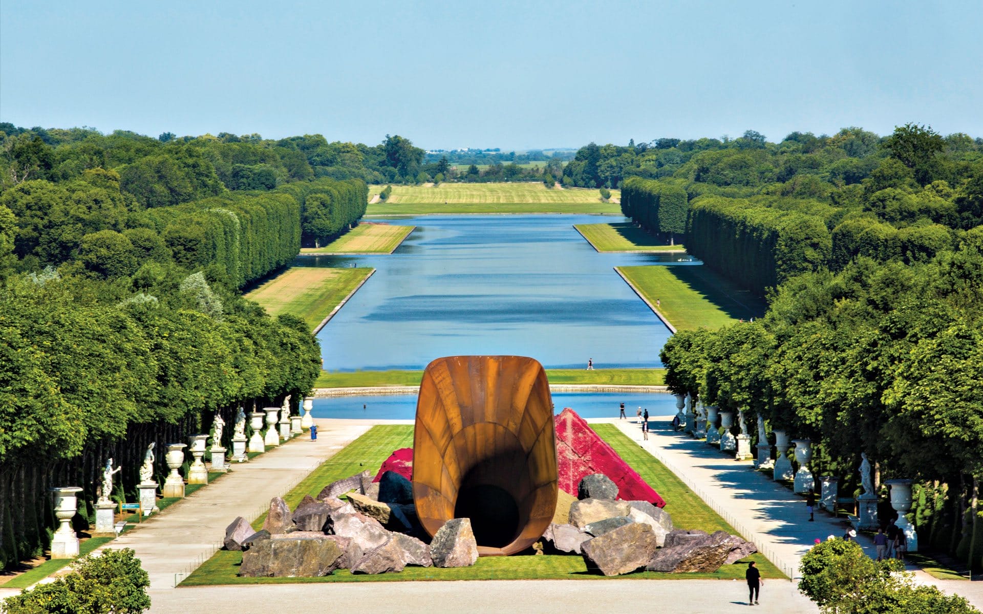 Sculpture in a garden with a long canal, surrounded by trees and statues, under a clear blue sky.