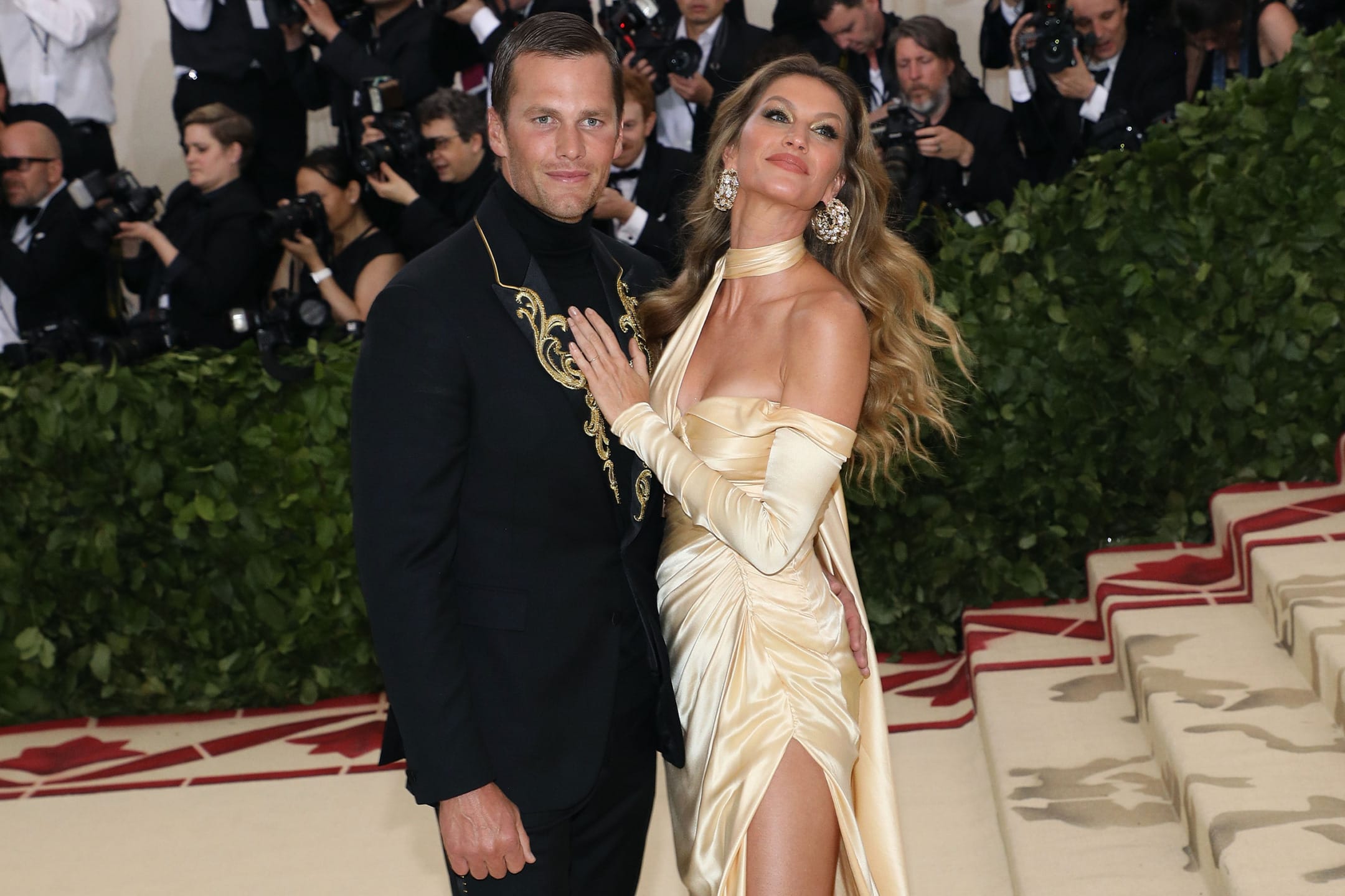 A couple posing elegantly on a staircase at a formal event with photographers in the background.