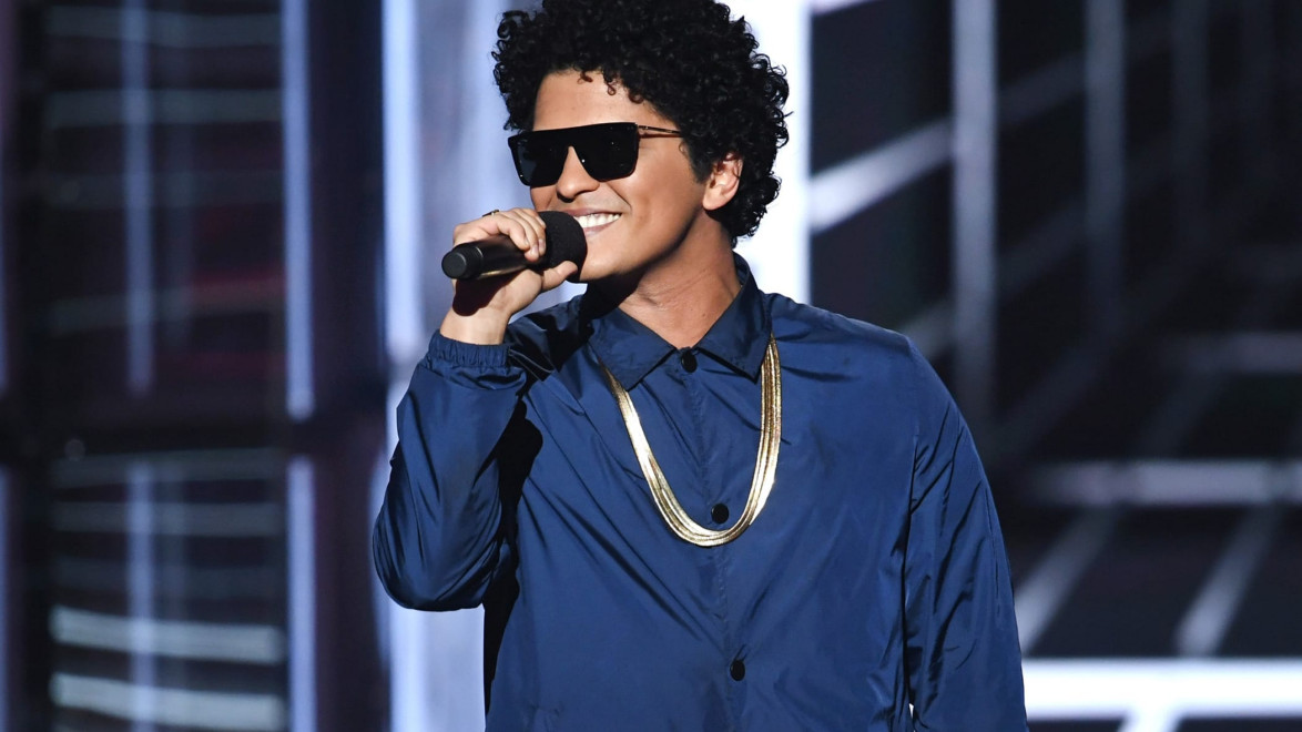 Singer wearing sunglasses and a blue jacket, holding a microphone on stage with a bright background.