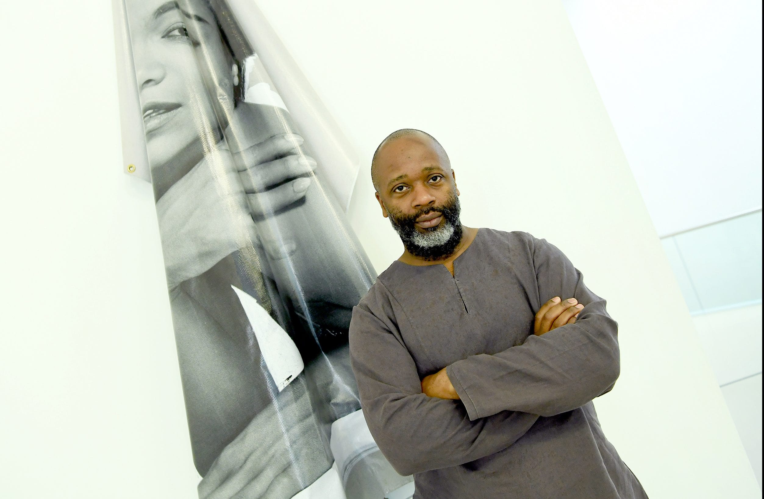 Man in dark clothing stands confidently with arms crossed next to a large monochrome art piece on a bright white wall
