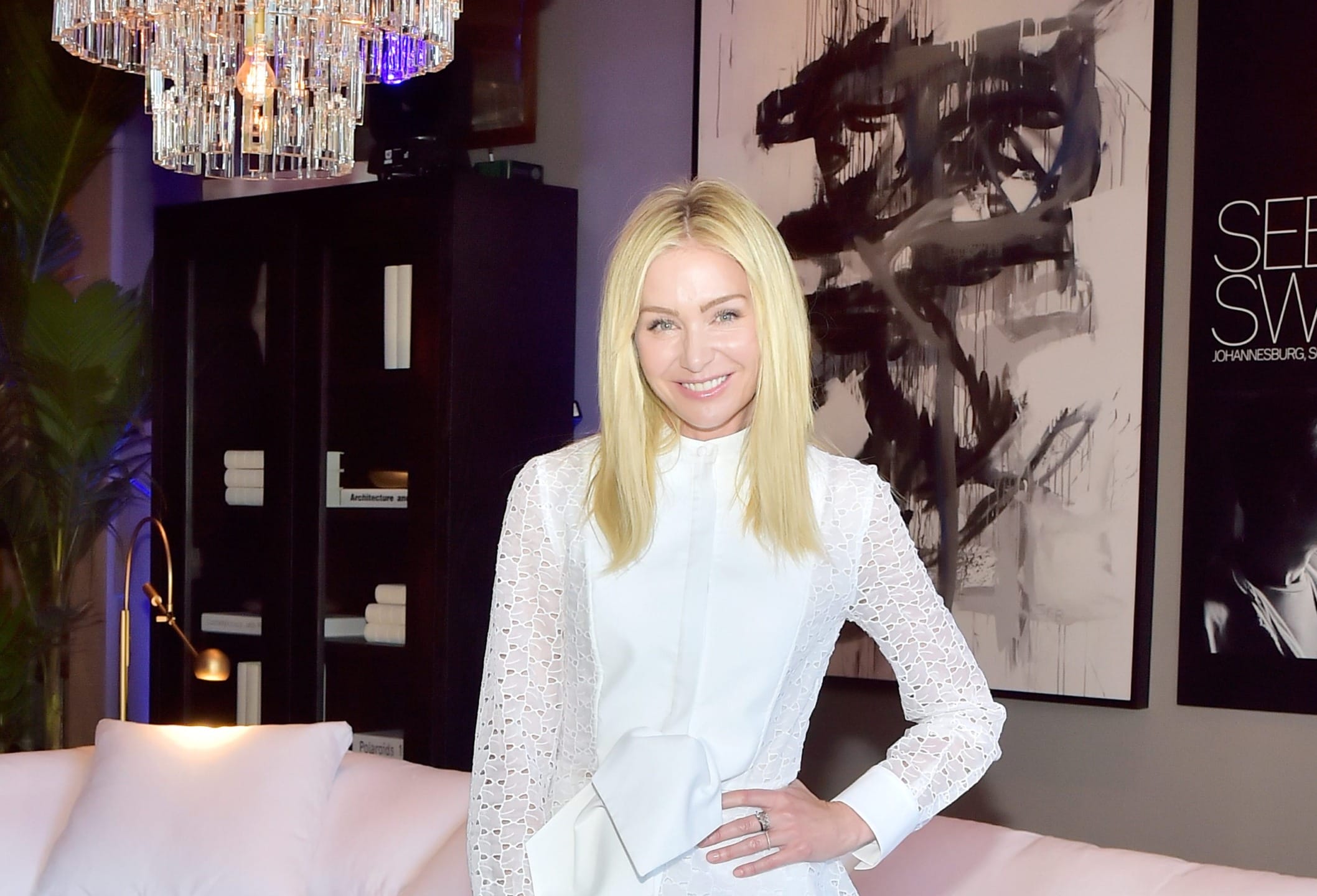 Person in a white dress standing in a stylish room with abstract art and a chandelier in the background.