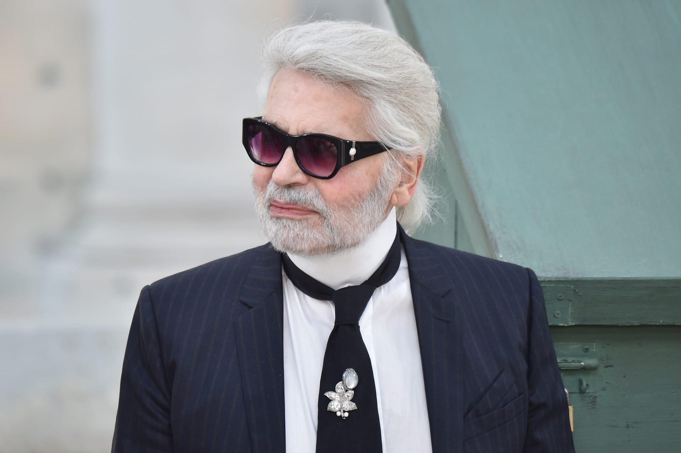 Man with white hair, sunglasses, and a suit with a tie, standing outside against a blurry background.