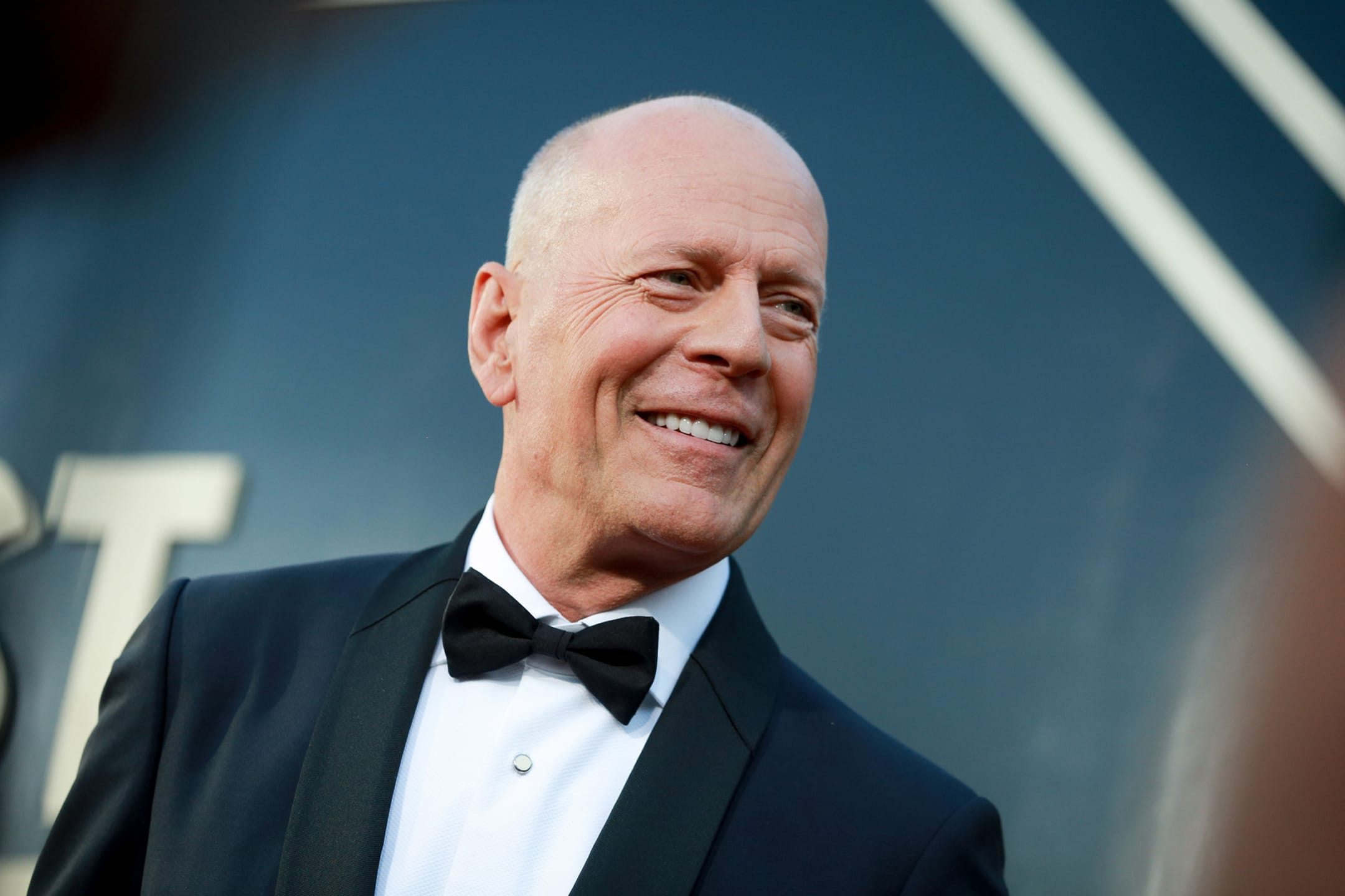 Man in a tuxedo smiling at an event with a dark background and white lines.