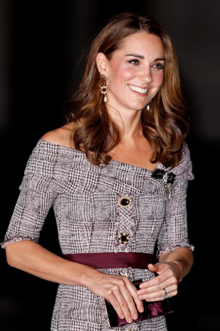 Smiling woman in an off-shoulder patterned dress holding a clutch, with brown hair and drop earrings.