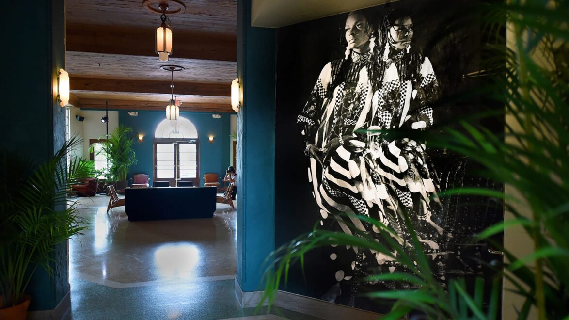 Hallway with large black and white artwork, plants, and retro lighting leading to seating area with arched window.