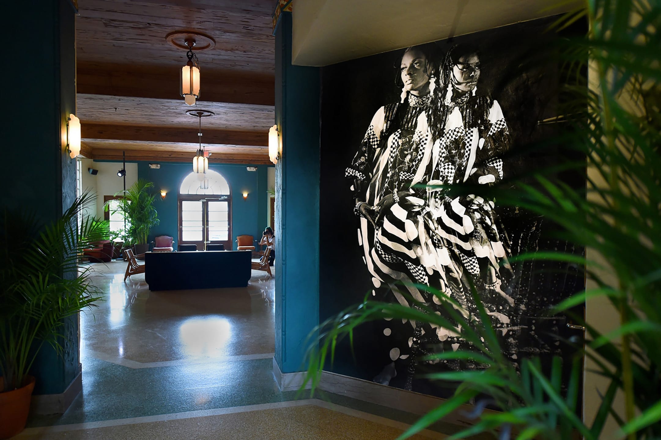 Hallway with large black and white artwork, plants, and retro lighting leading to seating area with arched window.