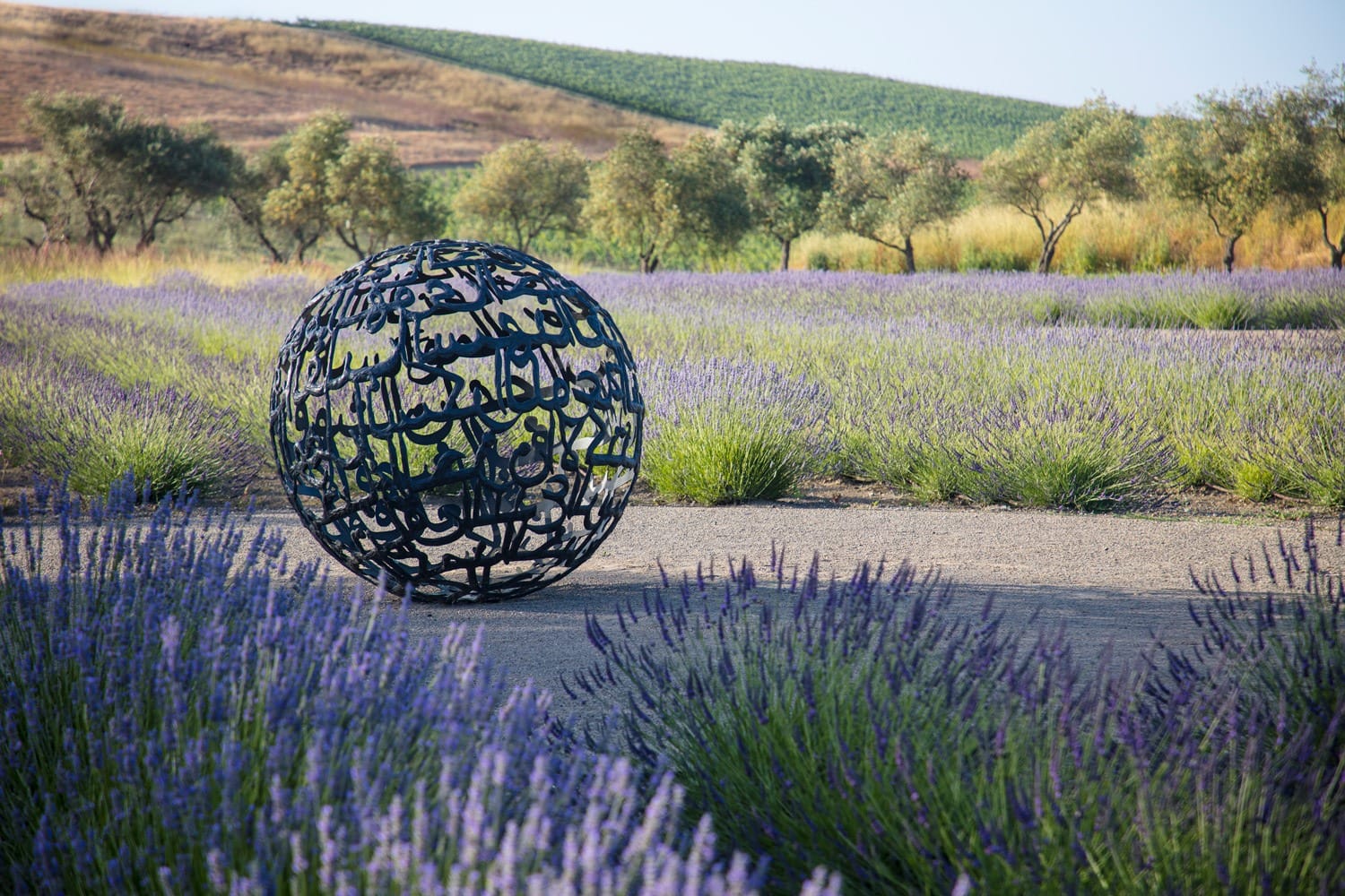 Sculpture with intricate design placed in a field of lavender, surrounded by olive trees and hills in the background.