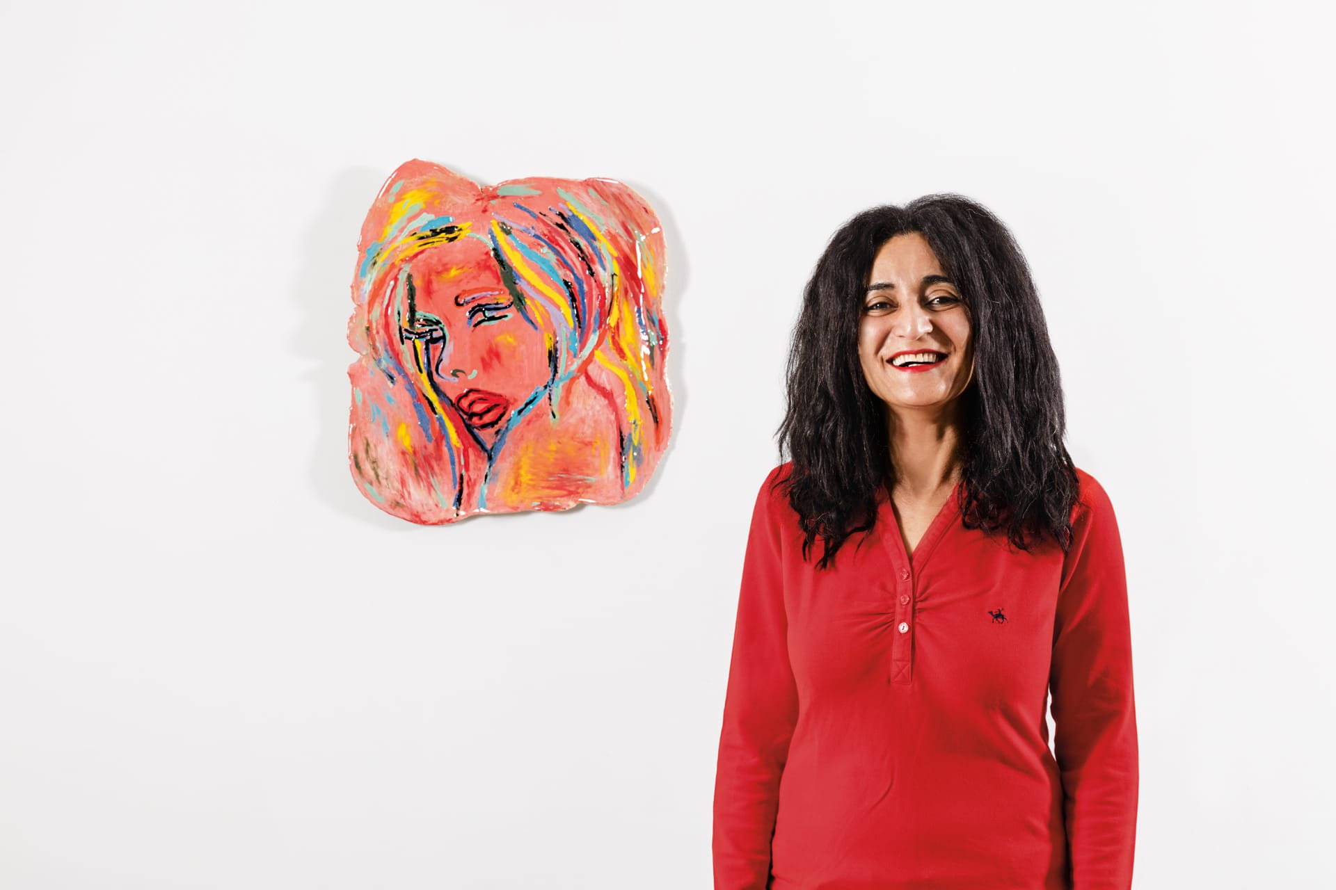 Woman in a red shirt smiling next to a colorful abstract painting on a white wall.