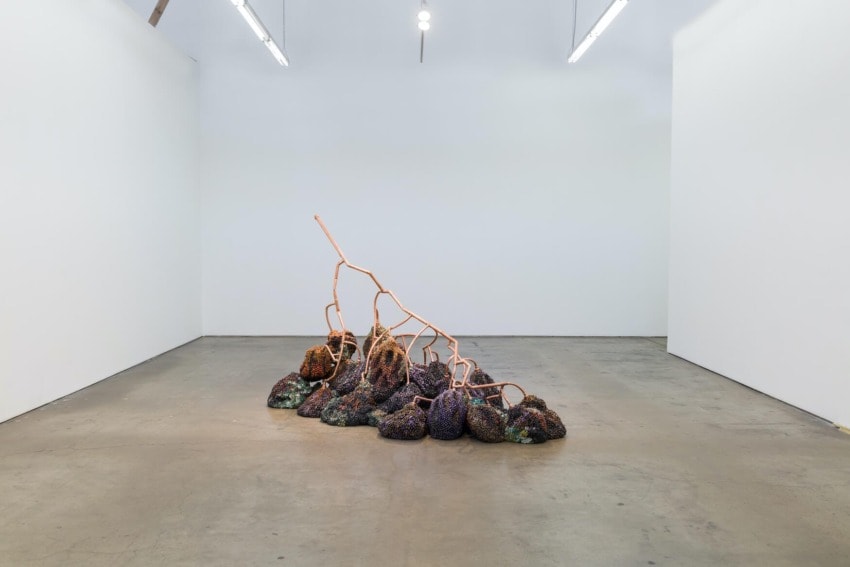 Contemporary art installation with copper pipes and textured multicolored materials on the floor in a minimalist gallery space.