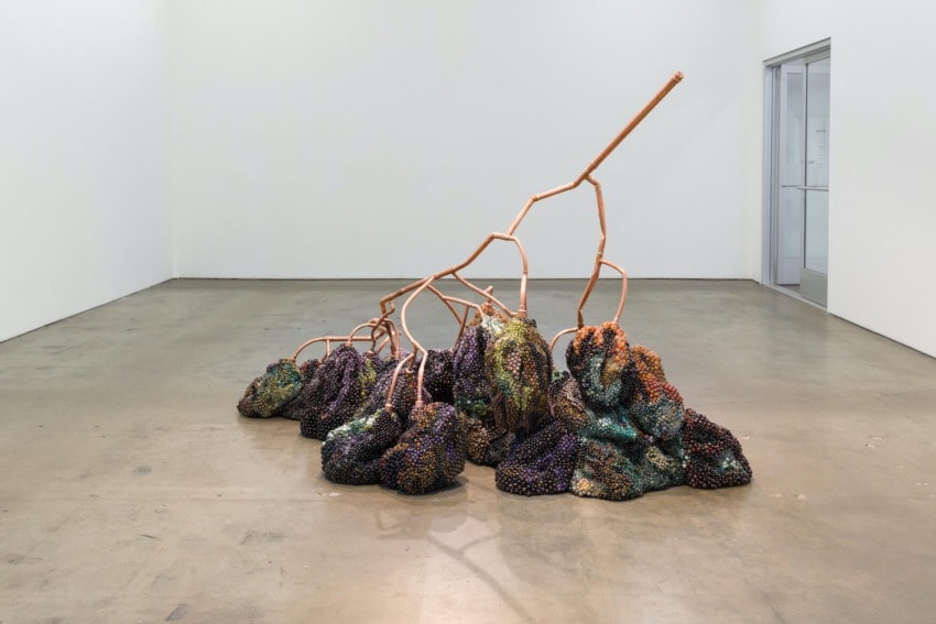 Abstract art sculpture with copper pipes and multicolored textured clusters on a concrete gallery floor.