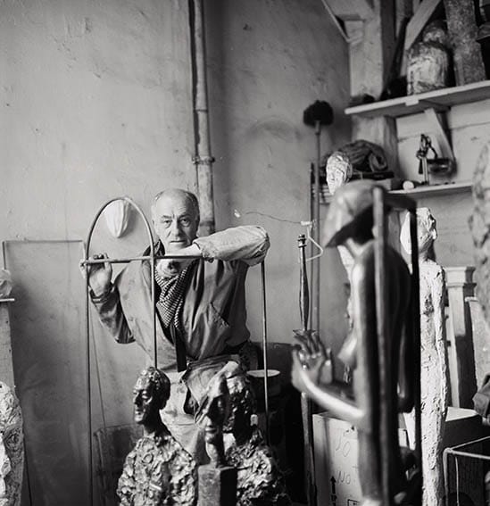Artist in a workshop surrounded by sculptures, working intently with tools and materials.